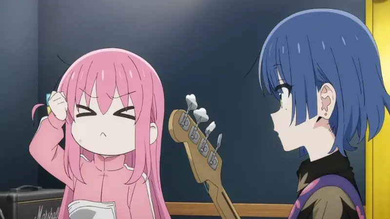 Hitori Gotoh looking panicked and stressed next to Ryo Yamada who is holding a bass guitar inside a dark recording studio.